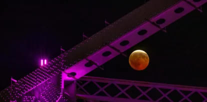 Un eclipse lunar parcial, visto a través de las vigas de acero -iluminadas de color morado- de Crescent City Connection, un par de puentes que cruzan el río Mississippi, en Nueva Orleans, el 27 de agosto de 2026. (AP Foto/Matthew Hinton)