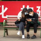Una pareja, con mascarillas para protegerse contra el coronavirus, mira un celular sentada en un banco delante de una pancarta que anima a la gente a usar tapabocas, en un parque público de Beijing, China, el 7 de mayo de 2020. (AP Photo/Mark Schiefelbein)